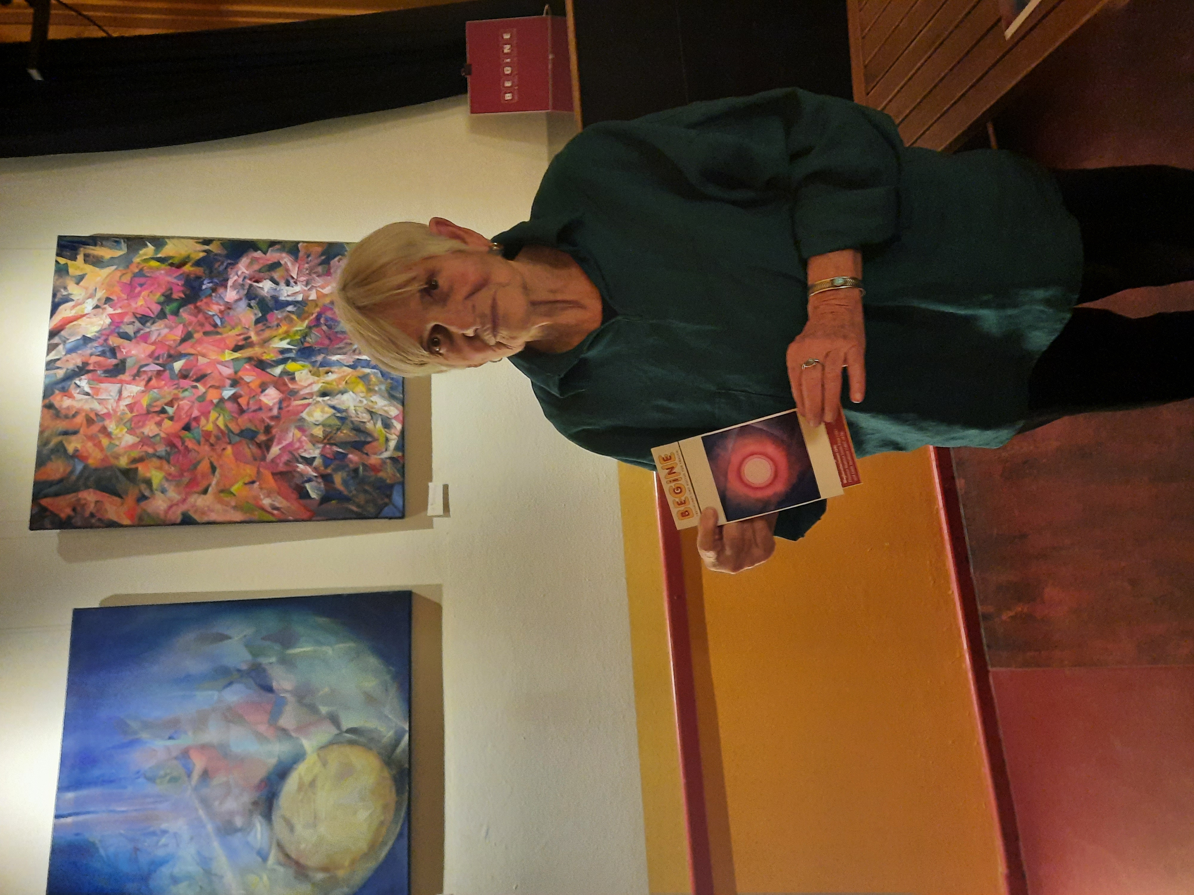 Ein Foto von einer älteren Frau, die steht und das BEGiNE-Programm in der Hand hält. Im Hintergrund hängen zwei von ihr gemalenen Bilder. 