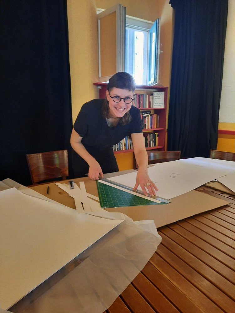 Eine junge Frau, die am Tisch arbeitet, ein Stück Papier schneidet und schaut uns an.