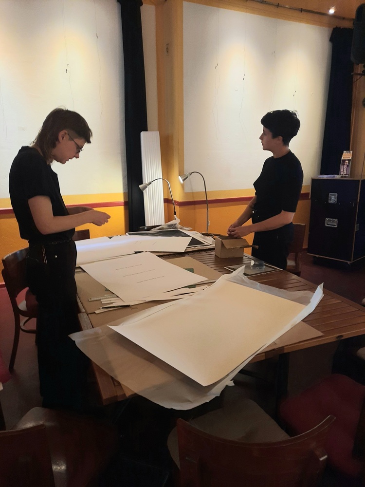 Zwei junge Frauen stehen im Raum am Tisch und vorbereiten eine Ausstellung 