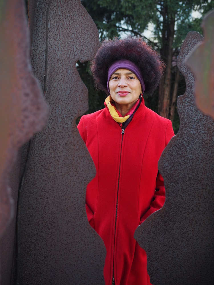 Ein Foto von einer Schwarzen Frau im roten Mantel, die hinter einer Skulptur steht. 