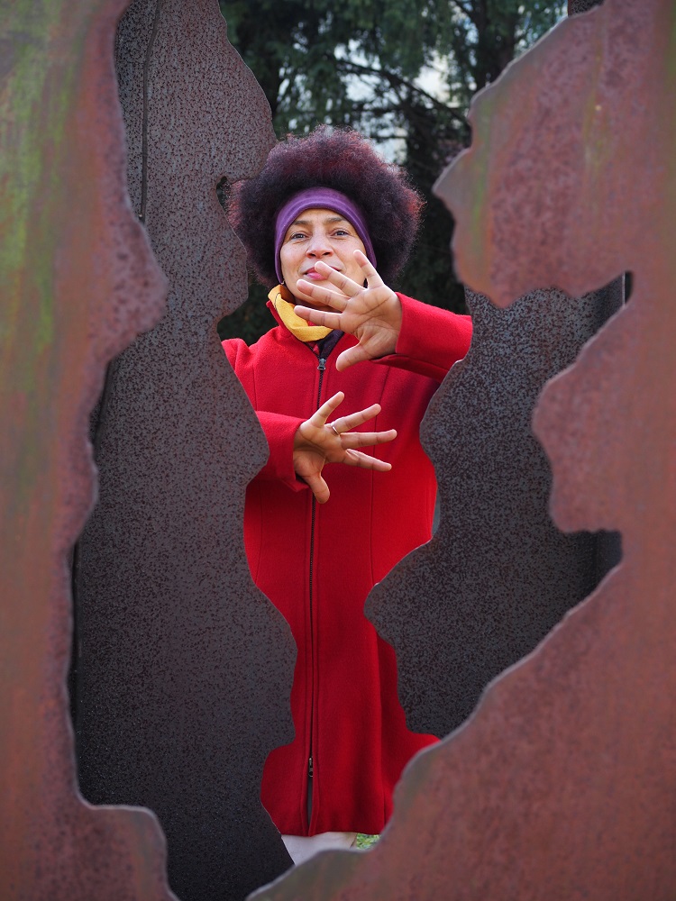 Ein Foto von einer Schwarzen Frau mit rotem Manten, die hinter einer Skulptur steht. 