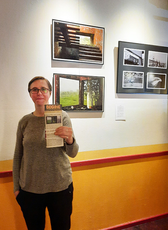 Ein Foto von einer Frau mit kurzem Haar und Brille, die vor hängenden Fotos auf einer Wand steht. Sie hält ineiner Hand ein Begine -Programm.