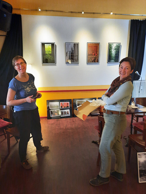 Ein Foto von zwei jungen Frauen, die vor einer Wand mit hängenden Fotos stehen, sich in unsere Richtung umdrehen und lächeln.