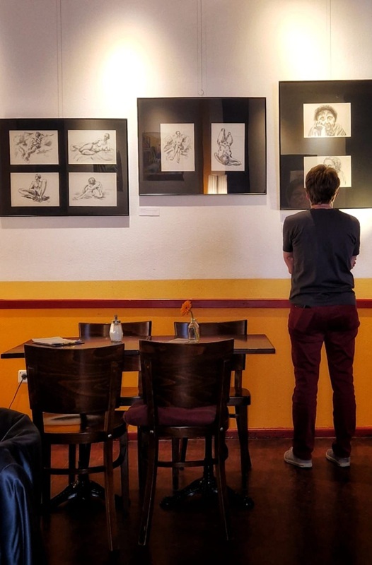 Ein foto von Bildern, die an der Wand hängen und einer Frau mit kurzem Haar, die vor diesen Bilder steht und sich die Bilder  anschaut. 