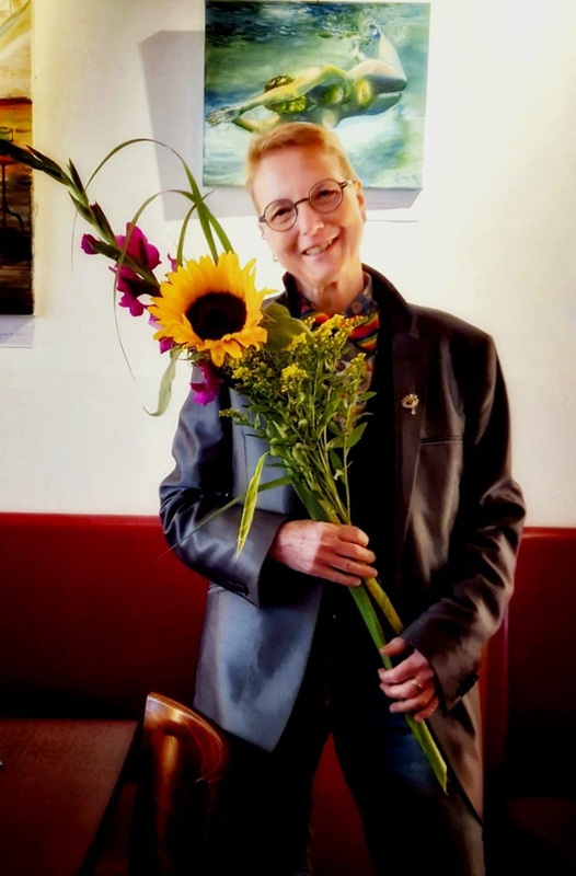 Ein Foto von einer Frau mit kurzem Haar, mit Sakko, einer Weste und einer Fliege, mit kurzem Haar und einer Brille, die viele Blumen in den Händen hält und lächelt.  