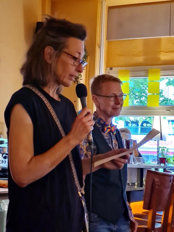 Ein Foto von einer Frau mit halblangem Haar, mit Brille, die einen Mikrofon in der Hand hält und liest etwas von einem Zettel, den sie in der anderen Hand hält. Hinter ihr steht eine andere Frau mit kurzem Haar und einer Brille. 