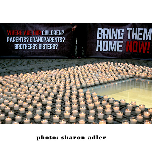 Ein Foto von vielen Kerzen, die auf dem Boden stehen. Im Hintergrung - Transparente mit Parolen: Bring them home. 